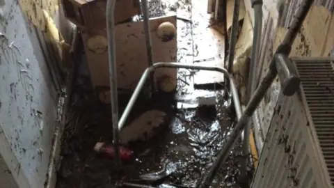 Paul Thomas The entryway of a home with thicj mud on the floor and up the walls, with various brooms and mops in the middle and a radiator on one side
