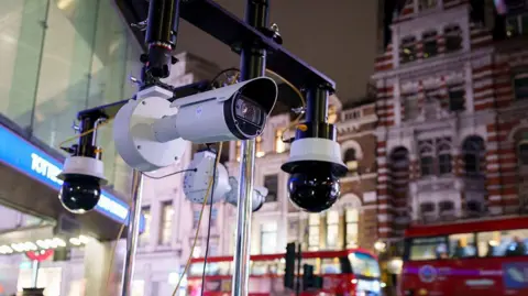 Close-up of security cameras mounted on a metal rig outside a London Underground station at night, with red double-decker buses and pedestrians in the blurred background.