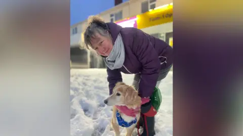 Simon Thake/BBC Lynn bends down next to a golden-colored dog. The dog is wearing a blue harness and a pink scarf around its neck, and it appears to be standing calmly on the snow-covered ground. Lynn is dressed in a dark purple winter coat and a light gray scarf. In the background, there is a building with a bright yellow sign that has some text on it.