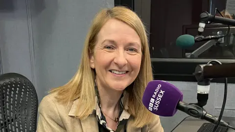 Katy Bourne sitting in a recording booth in front of a purple microphone which has "BBC RADIO SUSSEX" on it.