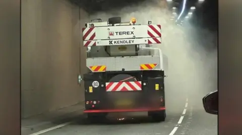 Eira D'Arcy Crane lorry with Terex Kendley company names on the white folded crane seen in the tunnel from the back with grey smoke coming from it and wing mirror of car behind is visible