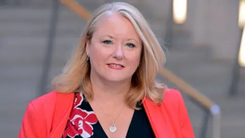 Getty Images A woman with blonde hair looks at the camera while walking in the Scottish Parliament. She is wearing a bright red jacket over a black top.