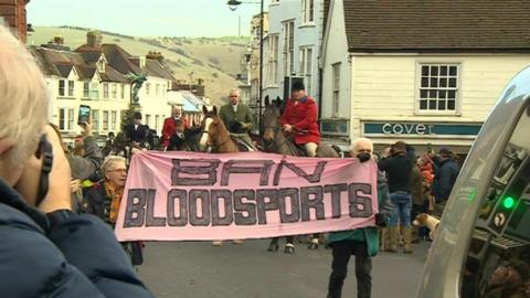 Lewes high street. Two demonstrators carry a sign saying 'Ban bloodsports' amid people on horses in hunting gear