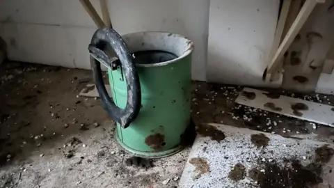 Andrew Turner/BBC A green tin toilet with the toilet seat broken, hanging from the top of the hinge mechanism Debris and dirt is on the floor nearby.