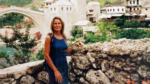 Ann is wearing a blue top and blue denim skirt and standing in front of the reconstructed Stari Most, also known as the Old Bridge in Mostar, Bosnia.