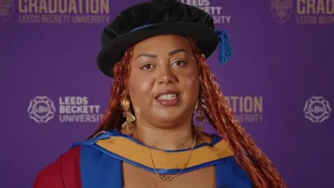 Emily Campbell, Olympic weightlifter, in a cap and gown as she receives an honorary doctorate. She stands in front of a purple background.