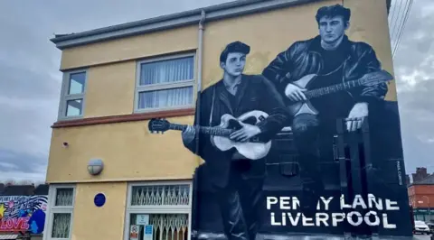 A black and white mural of Paul McCartney and John Lennon holding guitars sitting on a wall that reads Penny Lane Liverpool