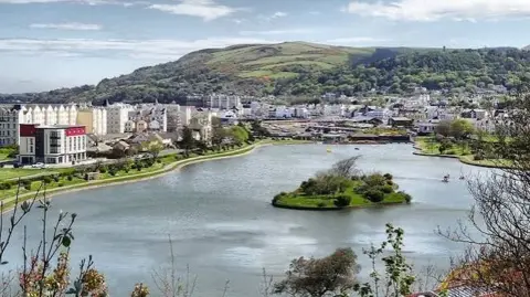 Mooragh Park in Ramsey, which has a large boating late with a small island in the middle. It has the promenade lined with large Victorian buildings and a hotel on the right, and the development within the town sits at the foot of the hills in the distance.