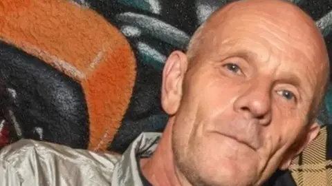 Paul Lumber looks at the camera. He is in his early 60s, white, bald and with light stubble. He is wearing a grey, shell-suit like top. 