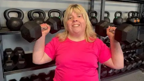 BBC Aisling McCooey holding two weights in the gym. She has blonde hair and a pink tshirt. She is smiling.