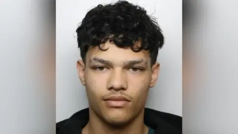 A mugshot of a man with short curly black hair and slits cut out of his eyebrows. He has a thin moustache and wears a black hoodie.