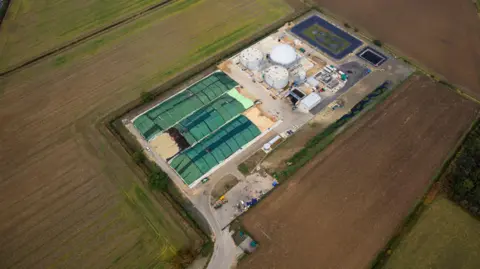 Pictured from above is a similar Future Biogas facility, Moor Bioenergy near Grantham which is comprised of a domed construction and two circular tanks with associated support buildings. The fields surrounding the facility are green with crops growing. 