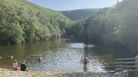 A number of people bathing in a rural reservoir surrounded by green hills. Old water extraction ironworks are visible from the right of the lake.