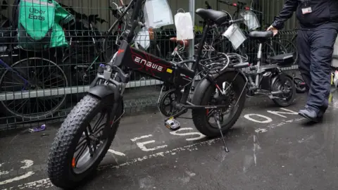 A modified e-bike, seized by police, is propped up by a kick-stand. There is a man behind wheeling a smaller e-bike and to the side opposite the camera there is a storage area containing a number of other bikes.