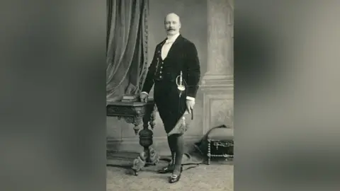 Geological Society of London Charles Dawson standing in a living room area. He is wearing a suit jacket, waistcoat, shorts, tights and black shoes with square buckle. He is holding a hat and has a sword tucked into his outfit. Charles has no hair and a moustache.