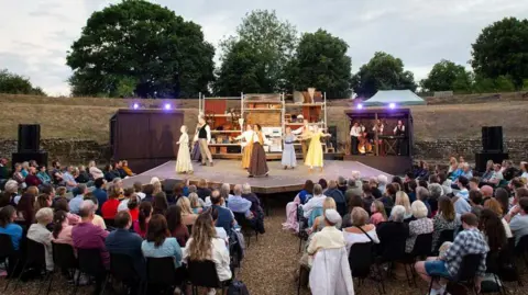 Elliot Franks An audience sit round the stage which juts out into the centre of the excavations at the Roman Theatre of Verulamium. There are eight actors standing on the stage and a band in a hut behind them upstage left. A set at the back of the stage consists of shelves with household items on them.