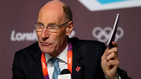 Getty Images A man in glasses and a dark suit sitting in front of a microphone. He is holding a booklet in his left hand
