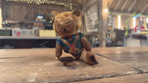 A tatty-looking World Cup Willy - a teddy bear of the 1966 mascot - sits on a wooden table in a workshop. He has closed eyes and is wearing a blue and red top.