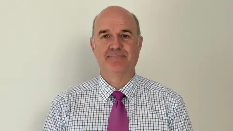 A bald man with grey hair on either side of his head wearing a white shirt with blue and red checked lines and a magenta tie standing in front of a plain white wall. 
