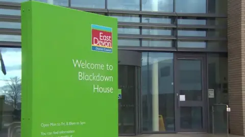 A lime green sign with the East Devon District Council logo, which is light blue, red and yellow. Below the logo it says welcome to Blackdown House and below on the left it states the opening hours. Behind the sign is the building's doors. 