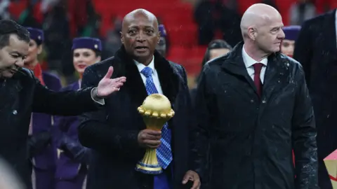 Caf president Patrice Motsepe, centre and dressed in a long black coat and white shirt and blue tie, carries the Africa Cup of Nations trophy in his right hand as he walks flanked by Fifa president Gianni Infantino.