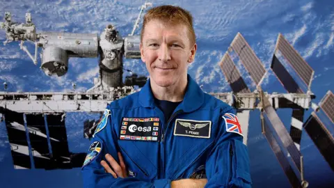 Getty Images Tim Peake, a man, wearing blue space overalls and crossing his arms, smiling. Behind him is an image of the International Space Station.