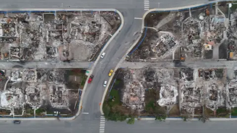 Several cars are parked on a street in between burned down houses.