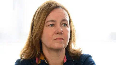 Getty Images Marie Goldman stands in front of a tree and greenery. She is wearing a navy blue top with floral detailing. She has shoulder-length brown hair and is smiling at the camera.