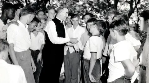 Boys Town Archives In an old black and white photograph, a priest in white shirt and black trousers, stands in the middle of a group of boys