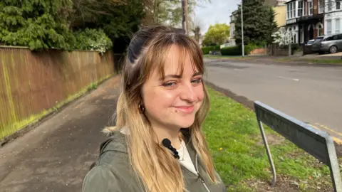 John Devine/BBC Nicole is smiling at the camera in a photo taken on a path at the side of the road. She has long, fair hair and is wearing a white top and khaki green jacket.