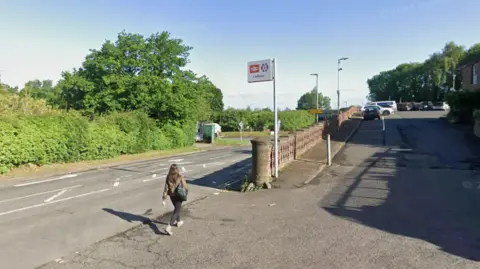 A woman walks besides an empty road on a sunny and clear day. A sign in front of her signals the entrance to Ledbury train station. 