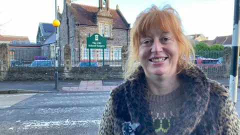 A smiling red haired woman, wearing a woolly leopard-print jacket, stands in front of a faded zebra crossing, with the sign for The Old School Surgery centred in the background.