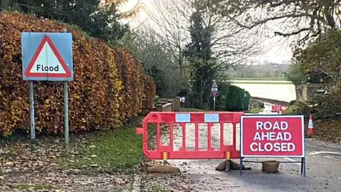 Radwell road closure showing a Road Ahead Closed sign and a large red barrier. You can see a flooded river in the background, flood sign, trees and bushes. 