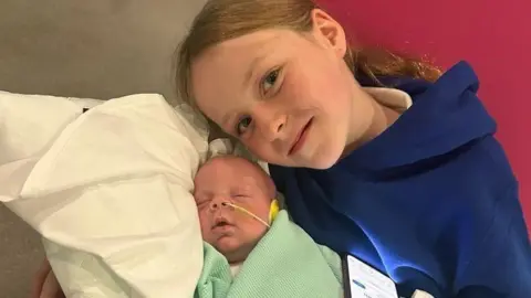 Chris Watt Photography Kobi is lying on a pillow in a green blanket. He has a feeding tube in his nose. His sister is wearing a blue hooded top. She is smiling at the camera and has her face near her baby brother. She has long brown hair.
