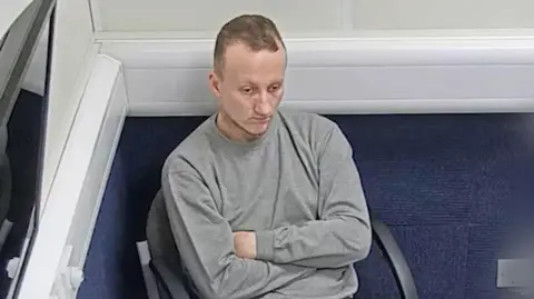 GMP Adam Thomson sits in a police interview room, wearing a grey sweatshirt, with his arms folded, staring impassively forwards.