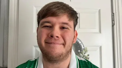 A smiling man is wearing a Celtic FC shirt, and is standing in front of a white door. He has short brown hair