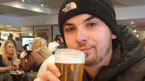 Liam Harvey wearing a black raincoat and a black North Face beanie hat. He is sitting in a restaurant holding a full pint of beer close to his face and smiling at the camera.
