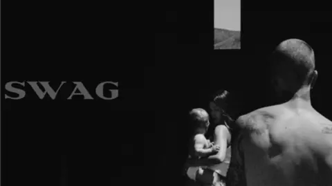 @lilbieber Justin Bieber stands with his back to the camera in a black and white image. Next to him is his wife Hailey Bieber holding a baby. The word "SWAG" is written on the right hand side of the image. Rolling hills are pictured in the distance.