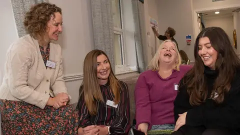 Four women sit on a sofa, laughing and smiling 