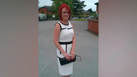 Tiffany Render has dyed-red hair and is smiling. She is standing near a building, dressed in a smart long white and black dress holding a black handbag. 