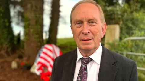 An older man with fair, receding hair. He is wearing a dark pinstripe suit with a white shirt and a purple paisley tie. He is standing in front of a field gate. Also behind him, out of focus, are some trees, and the memorial stone which is draped in the American flag.