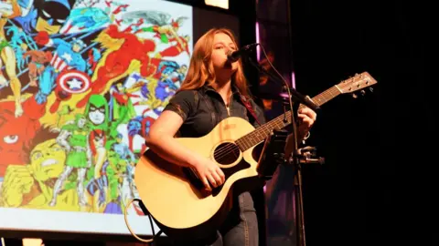 La Vida Liverpool & Jade Andreas Liv Jones, a young woman with long hair, stands singing and playing an acoustic guitar on a stage. 