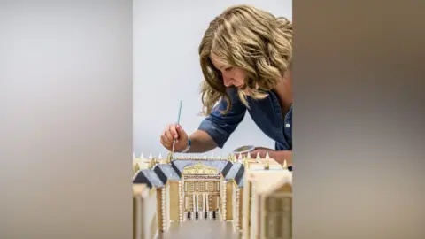 Eva Slusarek The Palace of Versailles made out of gingerbread. There is a woman with shoulder length hair looking at the gingerbread and holding what appears to be a thin paintbrush in order to add on the final touches to the project.
