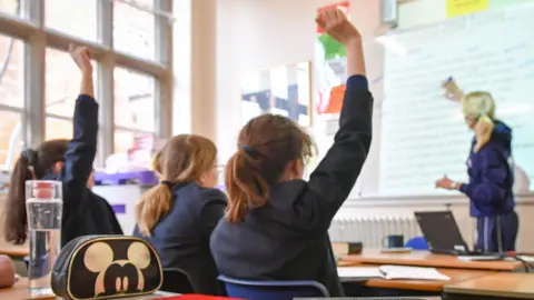 Murid media PA duduk di ruang kelas berseragam sekolah yang dihadapi guru di depan yang mengajukan pertanyaan kepada mereka dan dua dari mereka mengangkat tangan