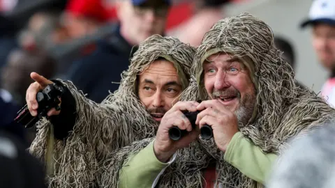 Getty Images Two men are pictured wearing military style camouflage. One is holding a pair of binoculars in both hands and grinning while the other man points something out to him. 