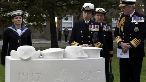 Royal Navy The Princess Royal, wearing a Royal Navy uniform, views a monument at Portsmouth Cathedral in the company of Royal Navy personnel. The white plinth is topped with four womens' service hats.