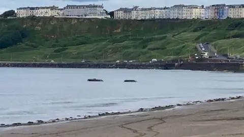Two cars stranded in the sea in Scarborough. There are cliffs in the background with a series of coloured houses on top.