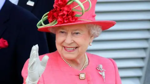 Getty The late Queen Elizabeth II wearing a pink outfit and a hat which has a flower design. One of her hands, in a white glove, can be seen waving in the picture. She is also wearing a silver broach. 