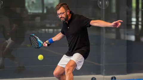 Bruby/Getty Images A man with a stern look about to hit a forehand with a padel racket, at an indoor centre. He has black hair, a black and grey beard, glasses and a necklace. He is wearing a black top and white shorts.