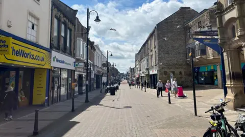Bishop Auckland high street. A number of shops and services line the street, including The Works and Newcastle Building Society. New Gate shopping centre is on the right.